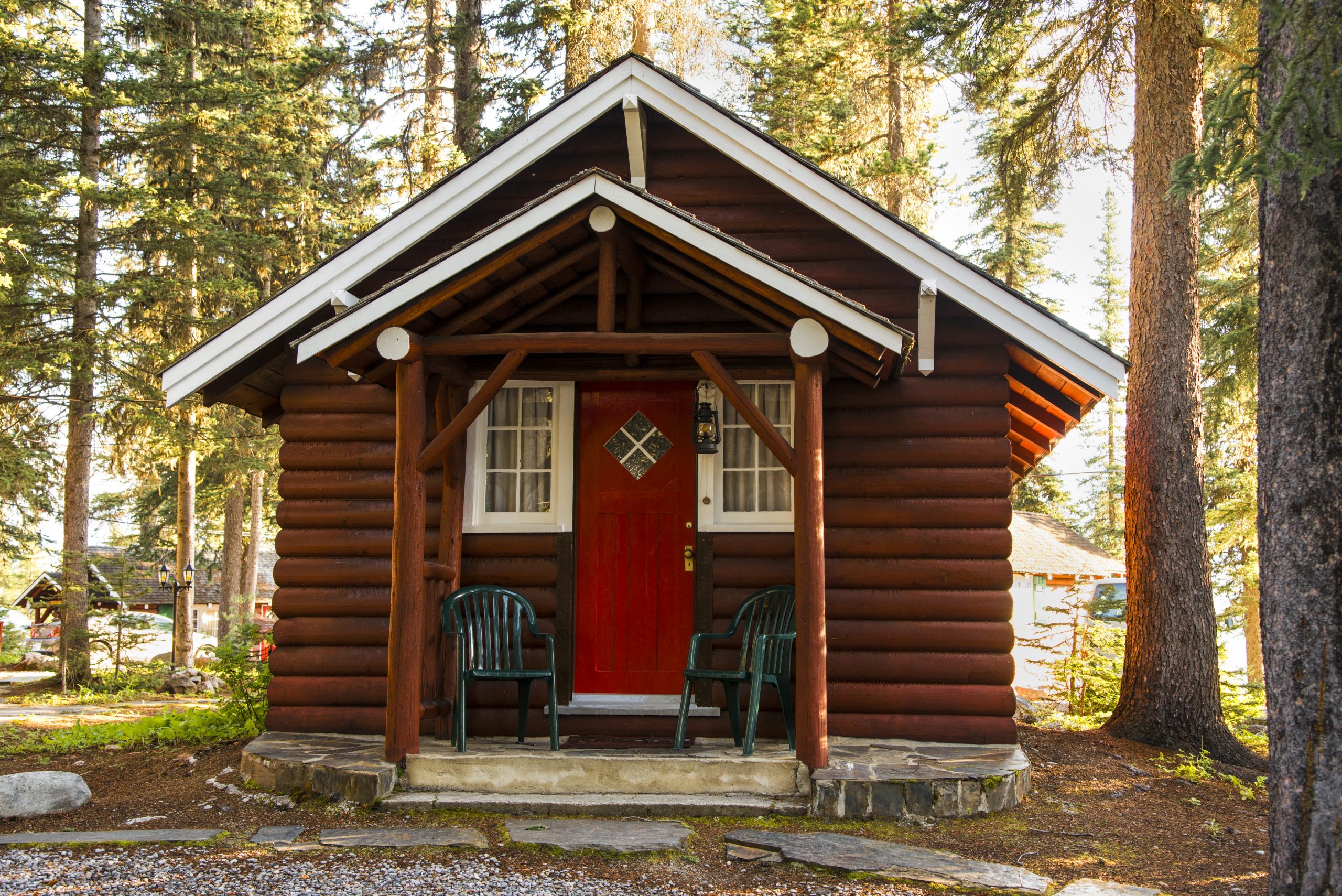 A small log cabin with a red door and two green chairs on the porch, surrounded by tall trees in a forest setting.