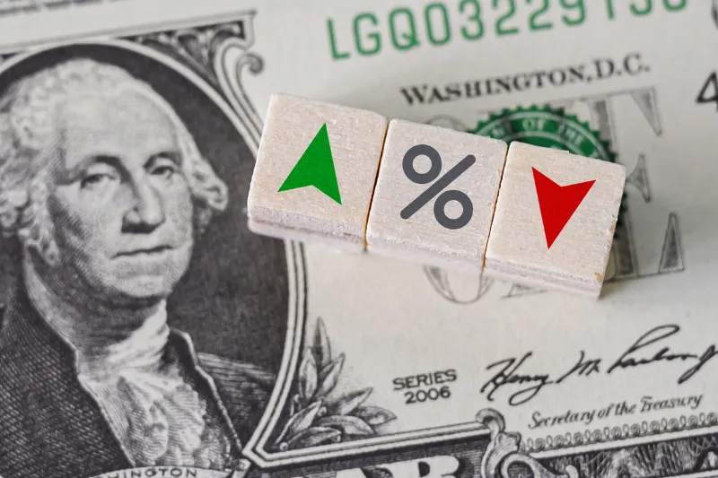 A close-up of a U.S. one-dollar bill with three wooden blocks on top displaying a green upward arrow, a percentage symbol, and a red downward arrow.